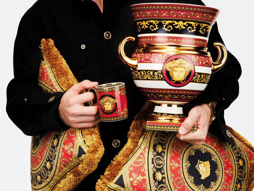 Porcelain design: A person is holding an intricately decorated red, black and gold porcelain vase and a matching cup featuring Baroque ornamentation and a Medusa motif.